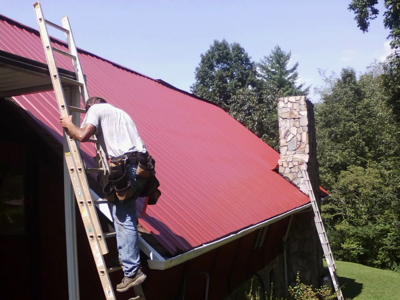 Roofer on a ladder beside a red metal roof and stone chimney — Soffit & Fascia Repair by Roofing Brothers in Kandiyohi